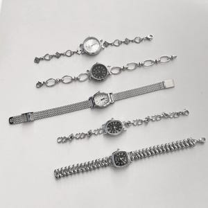 May include: Five silver-toned wristwatches with various face shapes and band designs are displayed on a white surface. The watch faces are round, oval, and square, with different dial colors and styles. The bands include chain links and mesh.