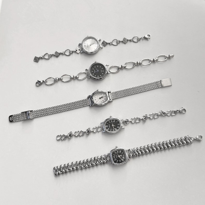 May include: Five silver-toned wristwatches with various face shapes and band designs are displayed on a white surface. The watch faces are round, oval, and square, with different dial colors and styles. The bands include chain links and mesh.