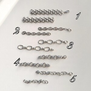 May include: An assortment of silver-colored chain links in various designs. The chains are displayed on a white surface, showcasing different styles, including oval, mariner, and curb chain variations. Each chain has a clasp for easy attachment.