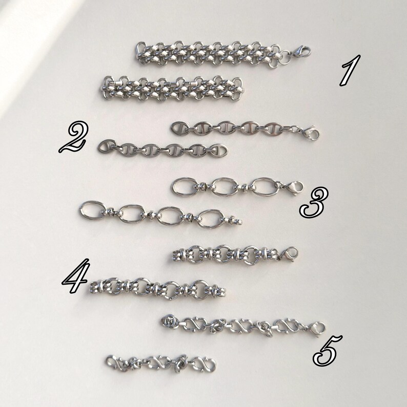 May include: An assortment of silver-colored chain links in various designs. The chains are displayed on a white surface, showcasing different styles, including oval, mariner, and curb chain variations. Each chain has a clasp for easy attachment.
