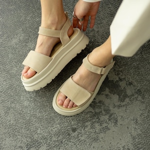 Könnte beinhalten: Beige Plateausandalen mit verstellbaren Riemen. Die Sandalen haben eine dicke, cremefarbene Sohle und ein oberes Material in Wildlederoptik. Das Fußbett ist für Komfort konturiert. Die Sandalen werden auf einer grauen Oberfläche gezeigt.