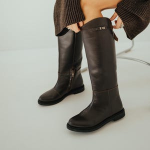 Puede incluir: Botas altas de cuero marrón oscuro con suela plana y cremallera lateral. Las botas tienen un detalle de hebilla en la parte superior y están hechas de cuero liso y pulido. Un estilo clásico, adecuado para varios atuendos.