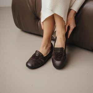 Pair of brown leather ballet flats with suede inserts, placed on a light gray surface, showcasing the sporty sneaker-inspired design and wide toe