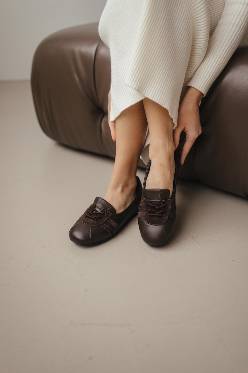 Pair of brown leather ballet flats with suede inserts, placed on a light gray surface, showcasing the sporty sneaker-inspired design and wide toe
