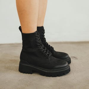 Puede incluir: Botas de combate negras con suela gruesa. Las botas tienen un diseño con cordones y una puntera redondeada. Las botas están hechas de un material similar al ante y tienen una lengüeta en la parte trasera del tobillo.