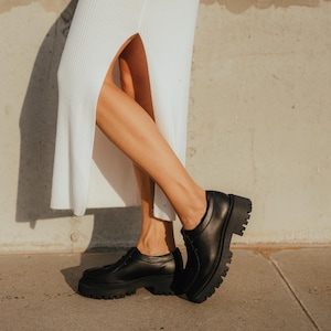 May include: A person wearing a white maxi dress with a side slit and black leather platform shoes.