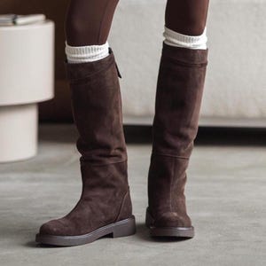 Puede incluir: Botas altas de ante marrón con suela plana. Las botas tienen un diseño ligeramente holgado. Los calcetines blancos asoman por la parte superior de las botas. Las botas son de un color marrón oscuro intenso.