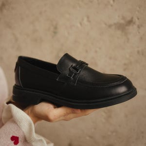 May include: A black leather loafer with a decorative buckle. The shoe has a rounded toe and a thick sole. The loafer is being held up by a person. The background is a neutral color.