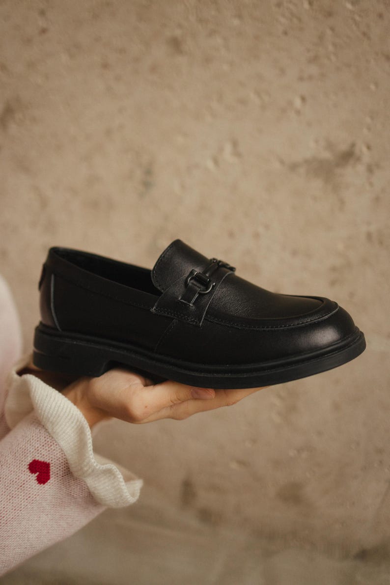 May include: A black leather loafer with a decorative buckle. The shoe has a rounded toe and a thick sole. The loafer is being held up by a person. The background is a neutral color.