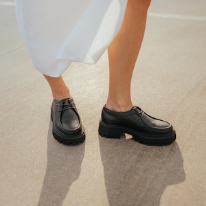 May include: A pair of black leather lace-up shoes with a chunky platform sole. The shoes are worn by a person wearing a white dress.
