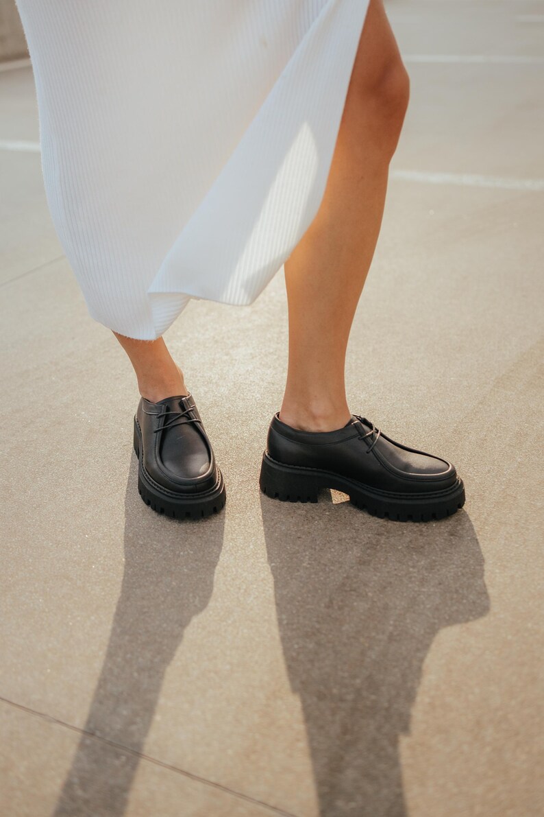 May include: A pair of black leather lace-up shoes with a chunky platform sole. The shoes are worn by a person wearing a white dress.