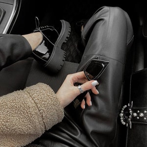 May include: Black leather trousers, a beige sherpa jacket, and black patent leather lace-up shoes with thick lug soles. A pair of black cat-eye sunglasses and a black handbag with a silver-toned pearl-embellished buckle are also visible.
