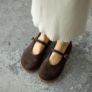 Könnte beinhalten: Ein Paar braune Mary Jane Schuhe aus Wildleder mit Riemen und Schnalle. Die Schuhe haben eine runde Spitze und eine hellbraune Sohle. Getragen von jemandem, der ein cremefarbenes Kleidungsstück trägt.