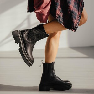 May include: A pair of black leather ankle boots with a chunky heel and a zipper on the side. The boots are worn by a person with one leg raised, showing the sole of the boot.