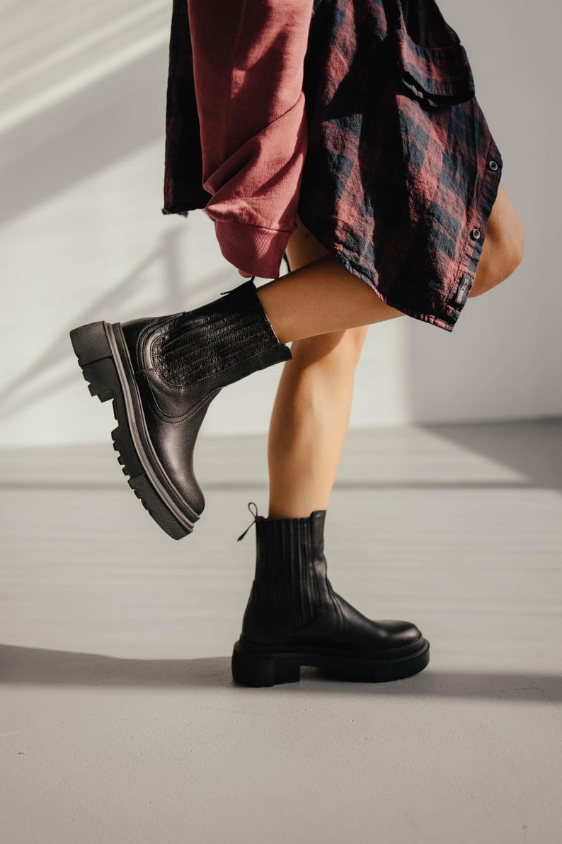 May include: A pair of black leather ankle boots with a chunky heel and a zipper on the side. The boots are worn by a person with one leg raised, showing the sole of the boot.