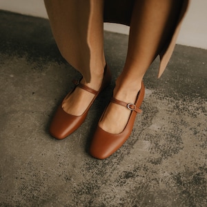 May include: A pair of brown leather shoes with a buckle strap. The shoes have a square toe and a low heel.