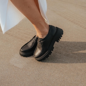May include: Black leather lace-up shoes with a chunky platform sole. The shoes are worn by a person standing on a concrete surface.