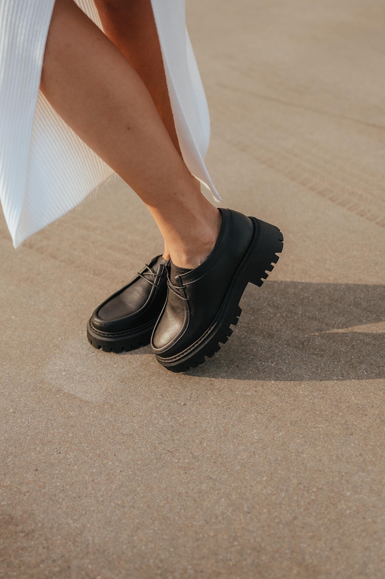 May include: Black leather lace-up shoes with a chunky platform sole. The shoes are worn by a person standing on a concrete surface.