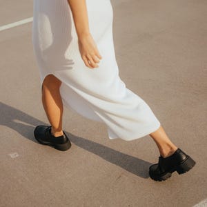 May include: A person wearing a white ribbed maxi dress and black chunky platform shoes walks on a concrete surface.