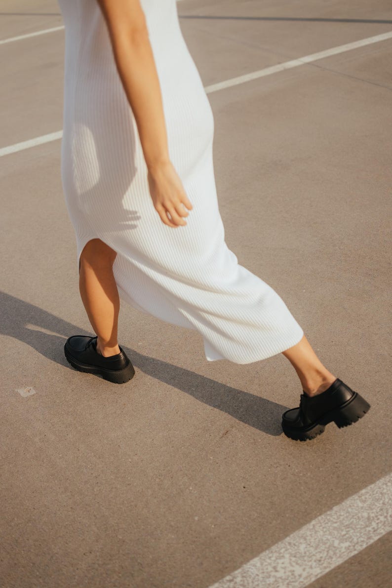 May include: A person wearing a white ribbed maxi dress and black chunky platform shoes walks on a concrete surface.