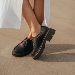May include: A pair of black leather lace-up shoes with thick white soles. The shoes have a chunky platform style and are worn by a person standing on a sandy surface.