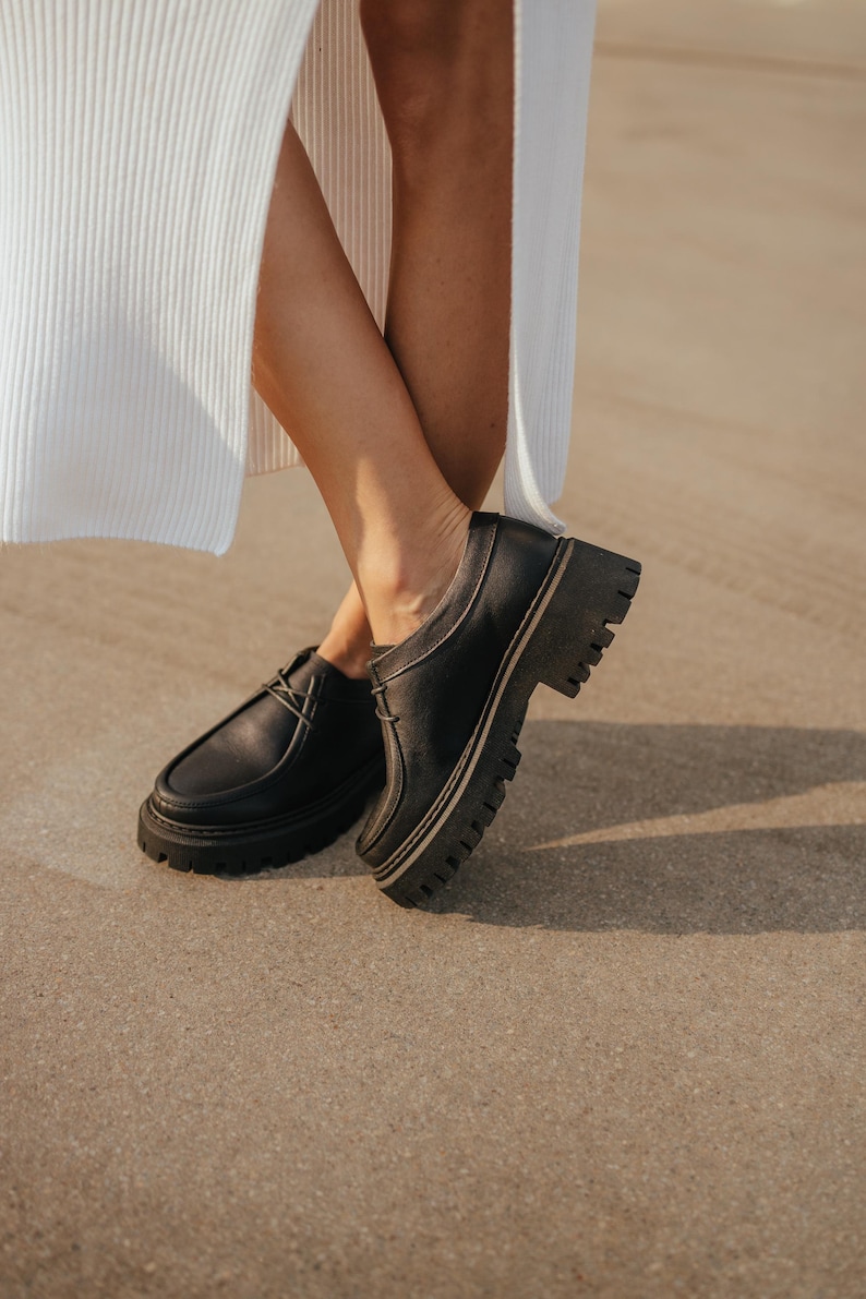 May include: A pair of black leather lace-up shoes with thick white soles. The shoes have a chunky platform style and are worn by a person standing on a sandy surface.