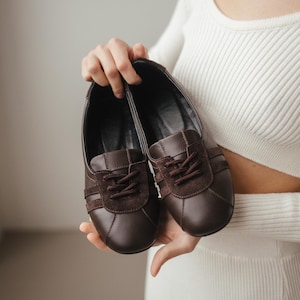 Person holding a pair of brown leather ballet flats with suede inserts, wearing a white ribbed sweater, minimalist background