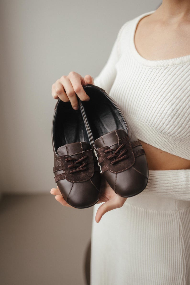 Person holding a pair of brown leather ballet flats with suede inserts, wearing a white ribbed sweater, minimalist background