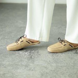 Puede incluir: Zapatos tipo mocasín de ante color beige con cordones y ribetes marrón oscuro. Los zapatos tienen un diseño clásico de mocasín con punta redondeada y tacón bajo. La imagen muestra los zapatos con pantalones blancos.