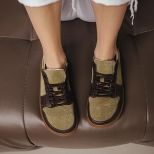 Close-up of handmade khaki leather and olive suede sneakers with decorative perforation on the toe, brown laces, and TEP sole, against a neutral background