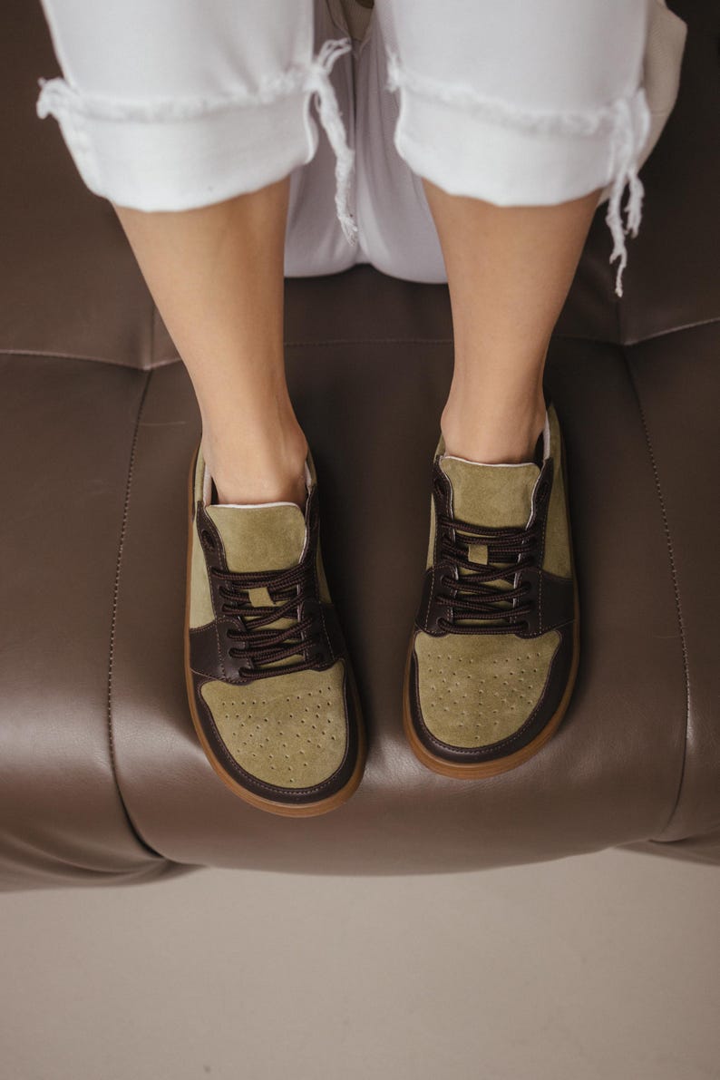 Close-up of handmade khaki leather and olive suede sneakers with decorative perforation on the toe, brown laces, and TEP sole, against a neutral background