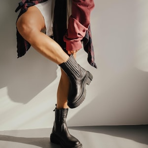 May include: A person wearing black leather ankle boots with a chunky heel. The boots have a zipper on the side and a pull tab on the back.