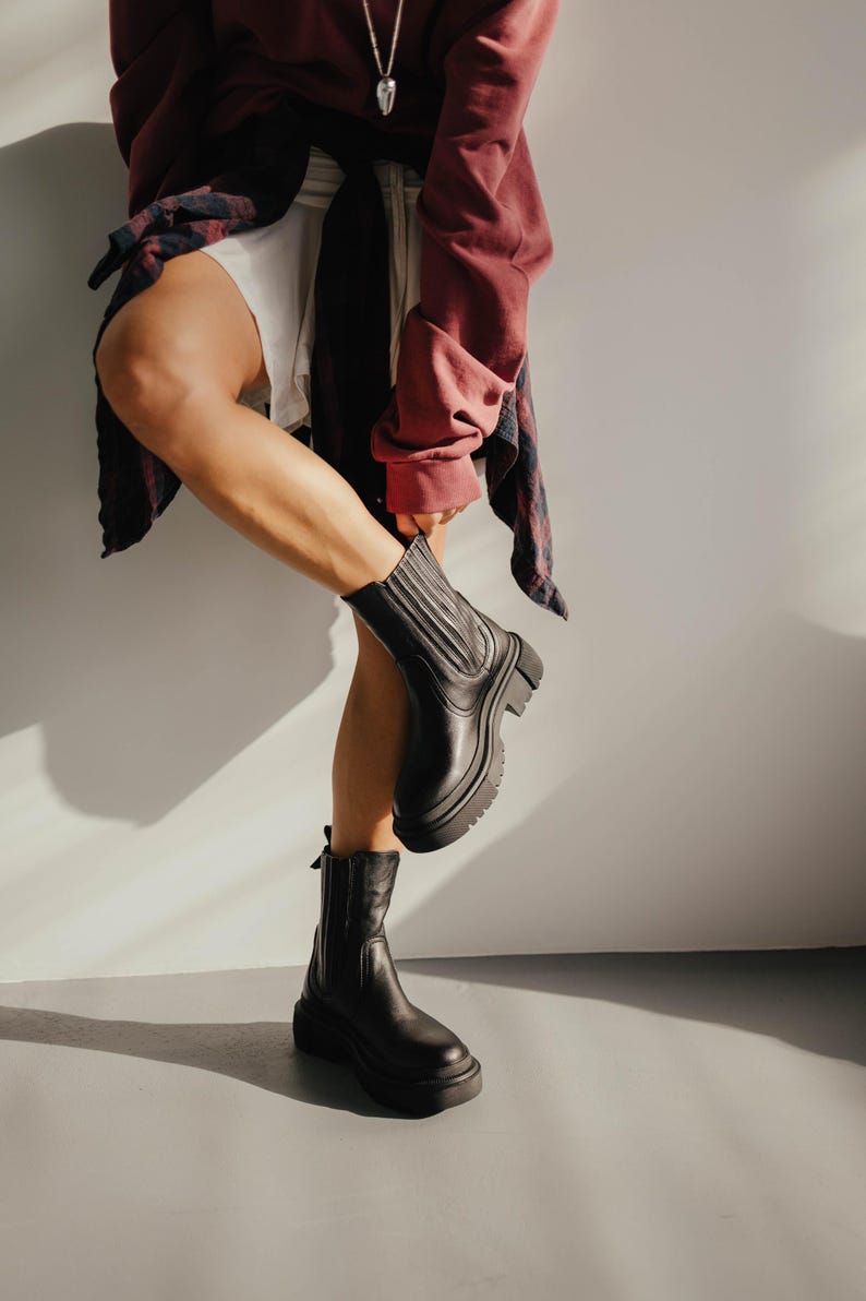 May include: A person wearing black leather ankle boots with a chunky heel. The boots have a zipper on the side and a pull tab on the back.