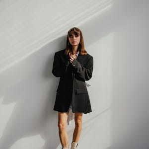 May include: A woman wearing a black blazer, a white shirt, and white boots stands in front of a white wall. The blazer has a long, loose fit and the boots have a chunky heel.