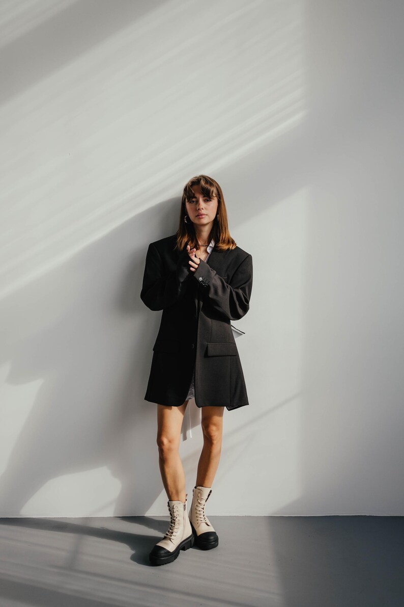 May include: A woman wearing a black blazer, a white shirt, and white boots stands in front of a white wall. The blazer has a long, loose fit and the boots have a chunky heel.