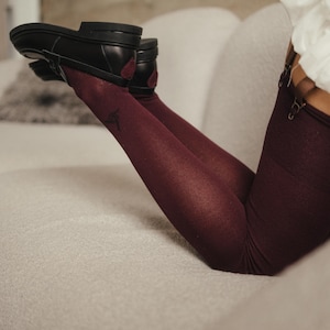 May include: Black leather shoes with a low heel and burgundy accents are paired with burgundy fishnet stockings. The stockings have a subtle pattern. The person is lounging on a light-colored sofa.