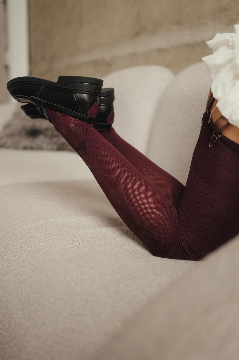 May include: Black leather shoes with a low heel and burgundy accents are paired with burgundy fishnet stockings. The stockings have a subtle pattern. The person is lounging on a light-colored sofa.