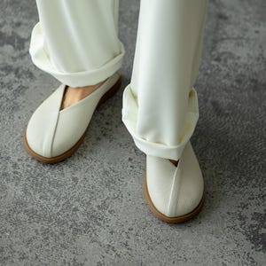 May include: Close-up of white slip-on shoes with a textured upper and a brown sole. The shoes are paired with white, cuffed trousers. The image is taken from a low angle, showing the shoes and the lower part of the legs.