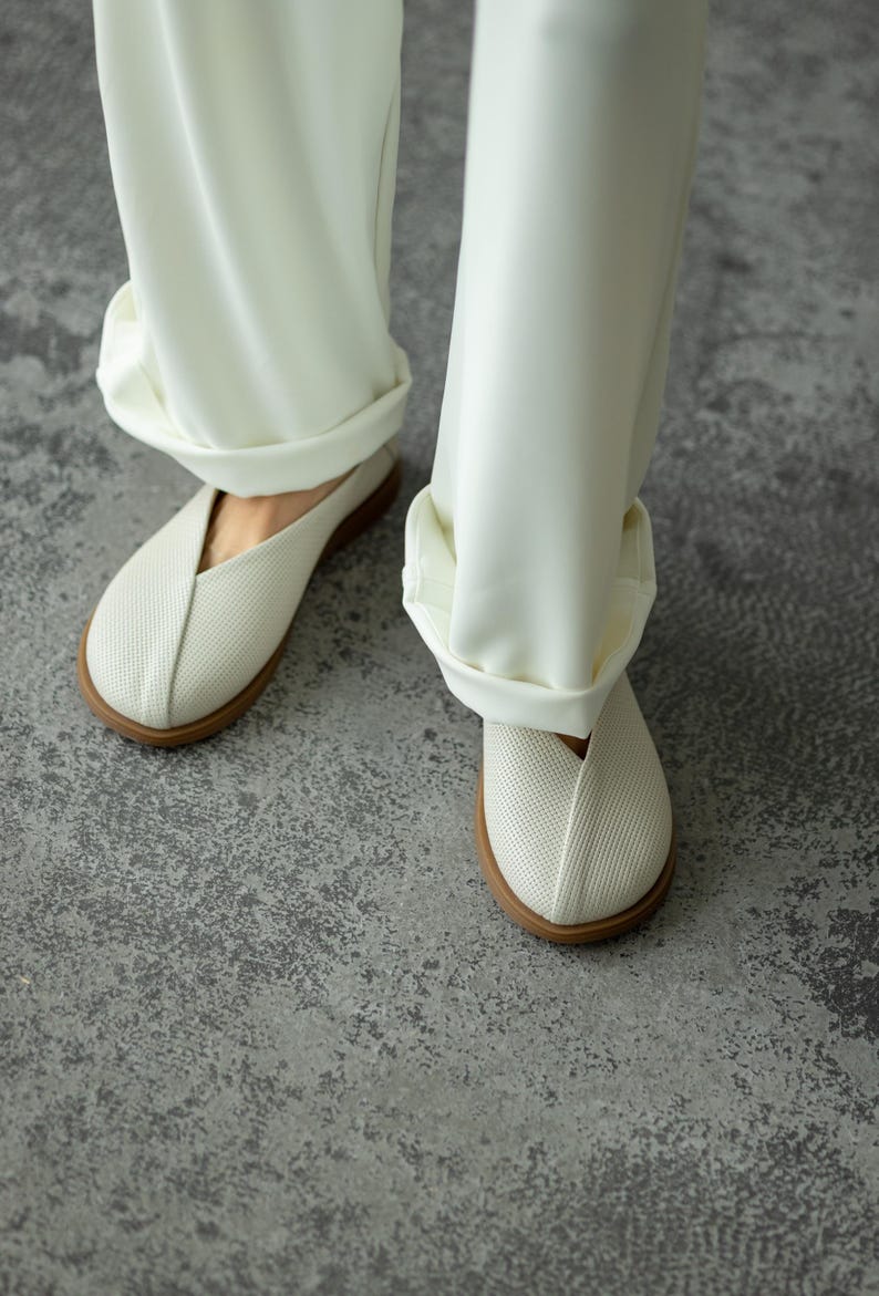 May include: Close-up of white slip-on shoes with a textured upper and a brown sole. The shoes are paired with white, cuffed trousers. The image is taken from a low angle, showing the shoes and the lower part of the legs.