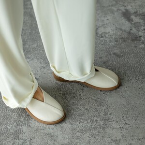 May include: Cream-coloured slip-on shoes with a perforated design and a brown sole. The shoes are paired with matching cream-coloured trousers. The image is taken from a low angle, showing the shoes and the lower part of the legs.