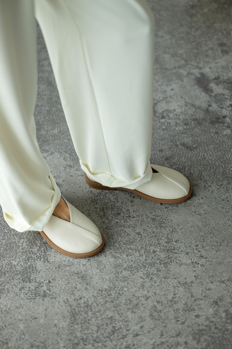 May include: Cream-coloured slip-on shoes with a perforated design and a brown sole. The shoes are paired with matching cream-coloured trousers. The image is taken from a low angle, showing the shoes and the lower part of the legs.