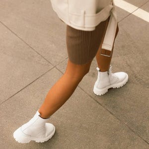 Puede incluir: Una persona con una chaqueta blanca y una falda marr&oacute;n, caminando con botas blancas de tobillo con suela gruesa.