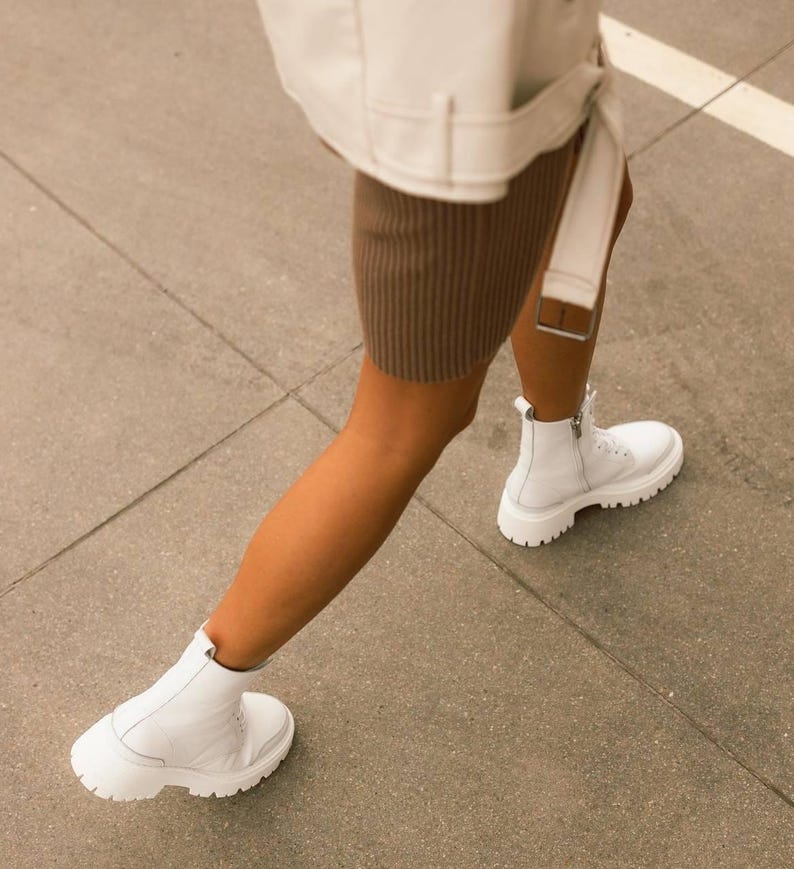 Puede incluir: Una persona con una chaqueta blanca y una falda marr&oacute;n, caminando con botas blancas de tobillo con suela gruesa.