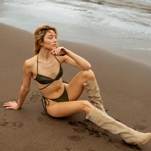 May include: A woman wearing a green bikini top and matching bottoms sits on a sandy beach. She is wearing tan knee-high boots and has her hand on her chin.
