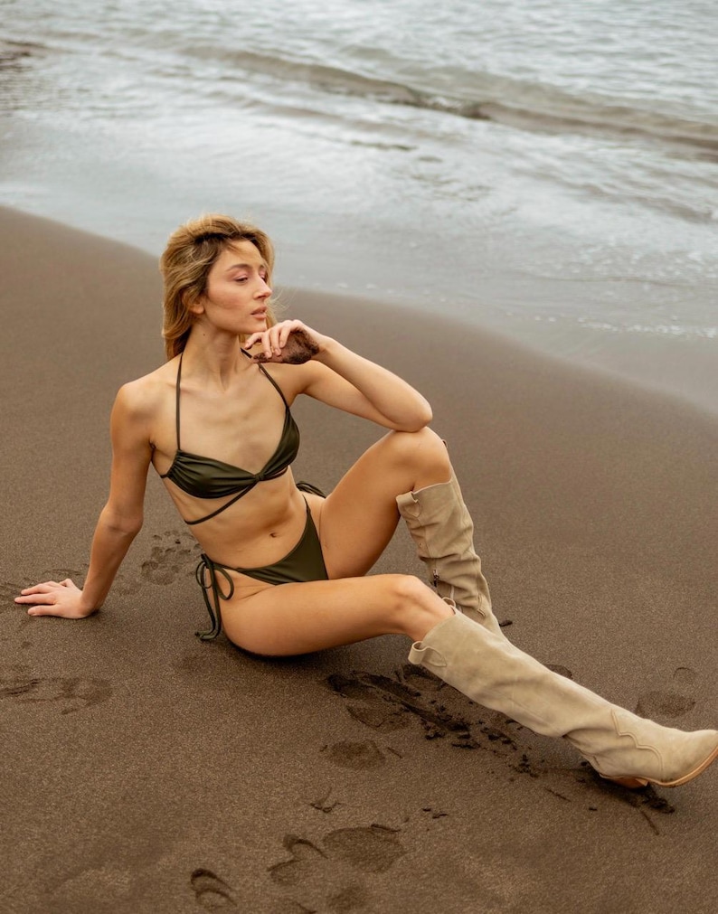 May include: A woman wearing a green bikini top and matching bottoms sits on a sandy beach. She is wearing tan knee-high boots and has her hand on her chin.