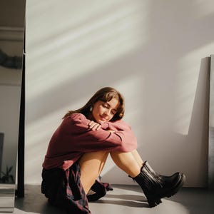 May include: A young person wearing a burgundy sweater and a plaid shirt sits with their legs crossed, wearing black ankle boots. The person is sitting on a white background.