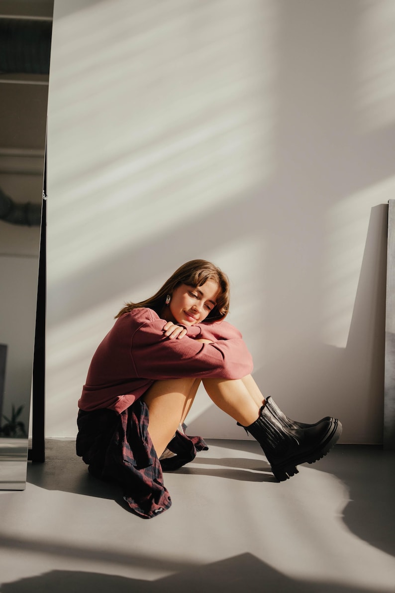May include: A young person wearing a burgundy sweater and a plaid shirt sits with their legs crossed, wearing black ankle boots. The person is sitting on a white background.