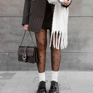 May include: A black pinstriped blazer, black sheer tights with a small heart pattern, white socks, and black patent leather lace-up shoes with thick lug soles. A black handbag with a silver chain strap and a white fringed scarf are also visible.