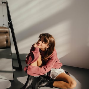 May include: A woman wearing a pink sweatshirt and black ankle boots sits on a gray floor in front of a mirror. She is smiling and looking to the side.