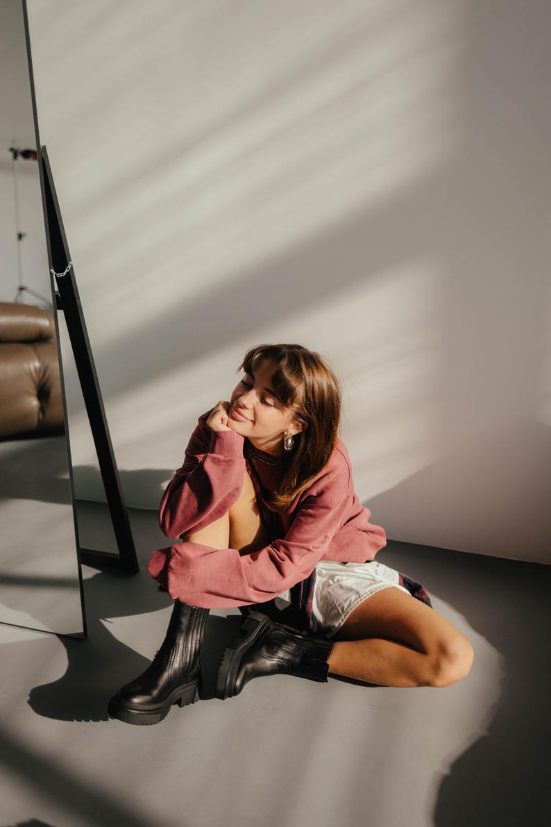 May include: A woman wearing a pink sweatshirt and black ankle boots sits on a gray floor in front of a mirror. She is smiling and looking to the side.
